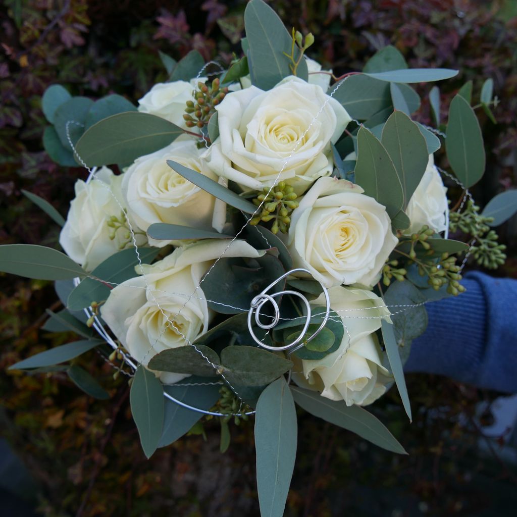 Brautstrauß aus weißen Rosen und grünen Blättern, gehalten von einer Person in blauem Pullover.