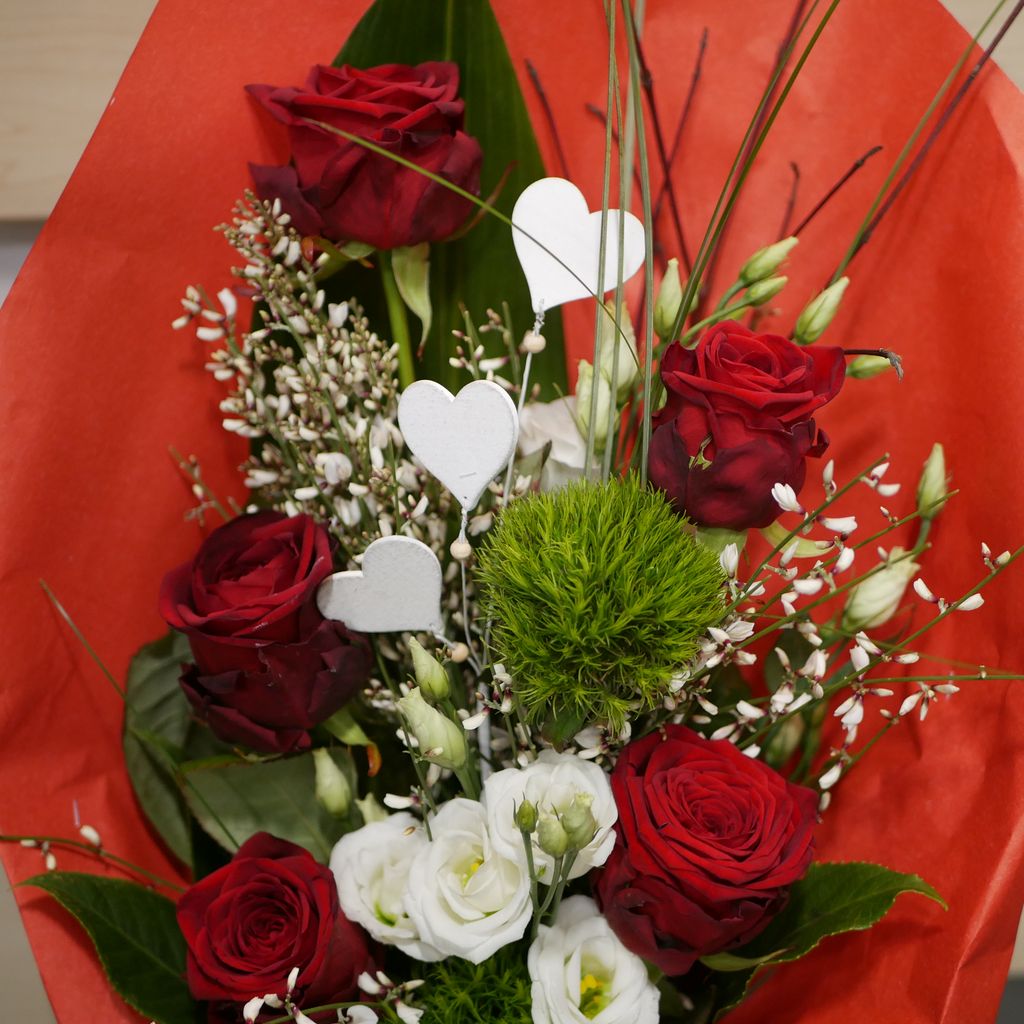 Roter Blumenstrauß mit roten Rosen, weißen Blüten, grünen Blättern und drei weißen Dekoherzen auf einem roten Hintergrund.