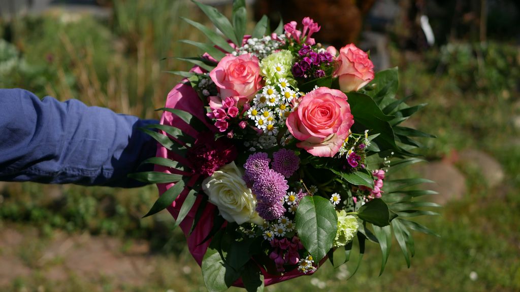 Ein Arm in blauem Hemd hält einen bunten Blumenstrauß mit rosa Rosen, weißen und lila Blumen sowie grünen Blättern in einem Garten.