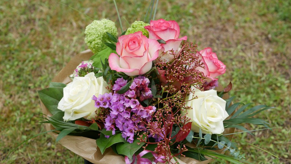 Bunter Blumenstrauß mit weißen und rosa Rosen, violetten Blüten und grünen Blättern vor einem unscharfen Gras-Hintergrund.