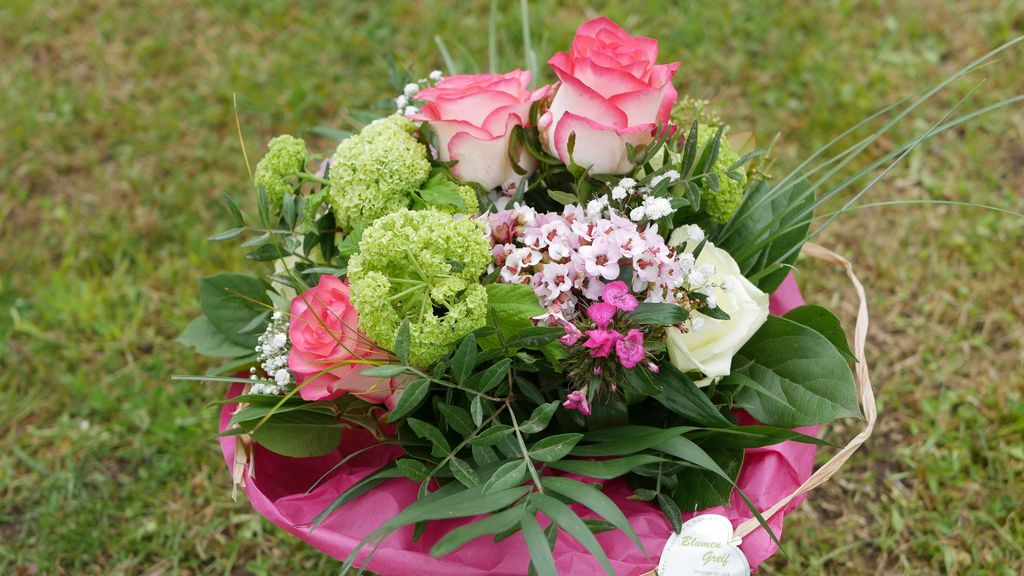 Bunter Blumenstrauß mit rosa und weißen Rosen, grünen Blüten und verschiedenen Blättern, präsentiert auf einem rosa Untergrund auf Gras.