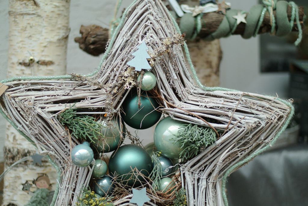 Ein dekorativer Stern aus verflochtenen Zweigen, geschmückt mit grünen Weihnachtskugeln, kleinen Tannenzweigen und Papiersternen, vor einem Hintergrund mit Birkenholzstämmen.