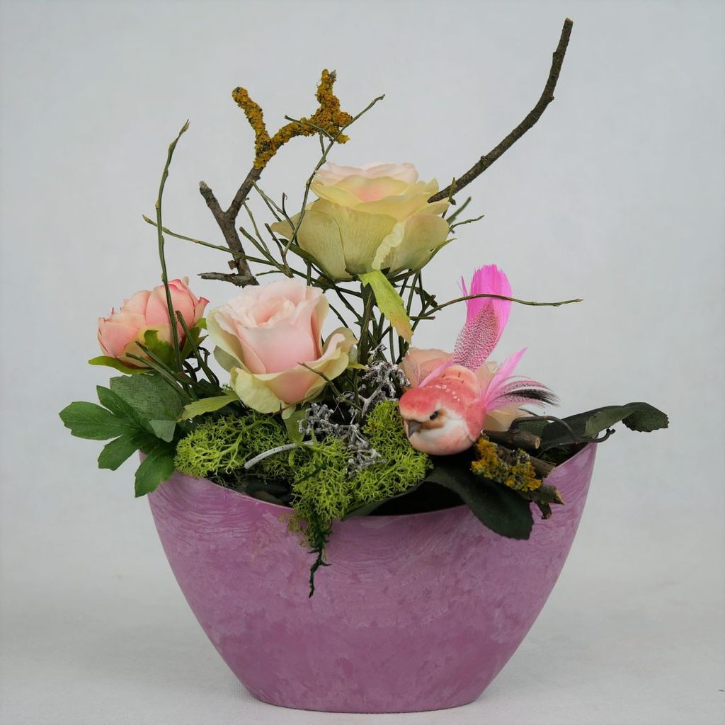 Blumenarrangement mit rosa und cremefarbenen Rosen, Zweigen, Moos und einem künstlichen pinken Vogel in einem violetten Übertopf.