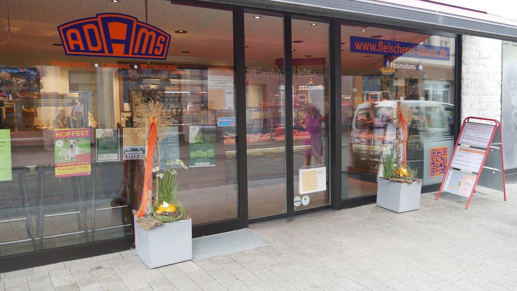 Glasfront eines Fleischerei-Geschäfts mit orange-blauem ADAMS-Logo, zwei Pflanzkübeln mit Dekoration und einem Aufsteller mit Informationen auf dem Gehweg.