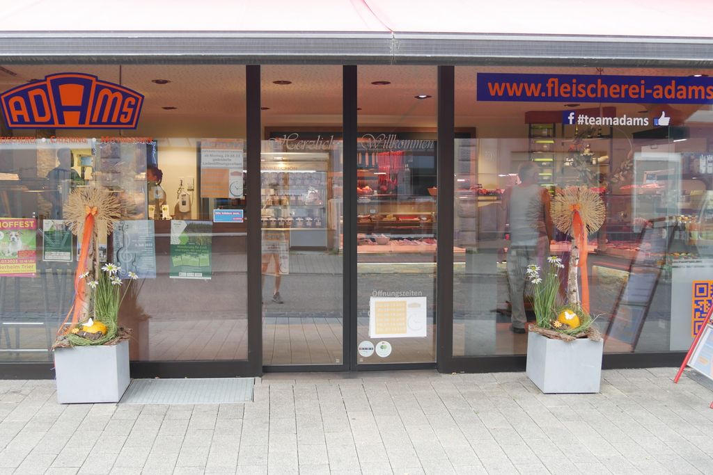 Eingangsbereich einer Fleischerei mit gläserner Schiebetür, Dekoration mit Blumen und Kürbissen links und rechts, dahinter Sicht auf Auslage mit Fleischwaren und eine Person im Laden.