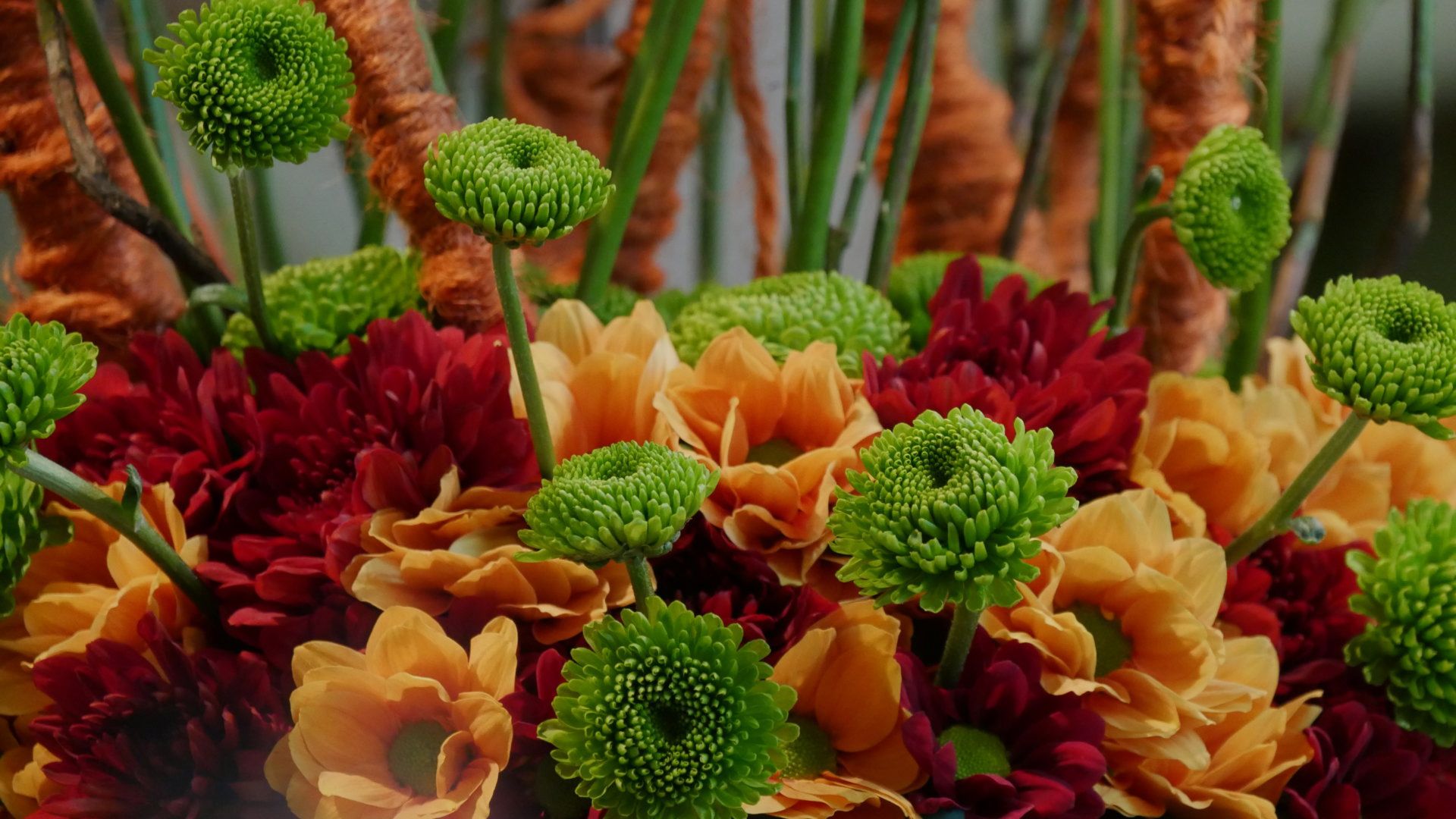 Nahaufnahme eines Blumenstraußes mit grünen Chrysanthemen, roten und orangefarbenen Blumen vor unscharfem Hintergrund.