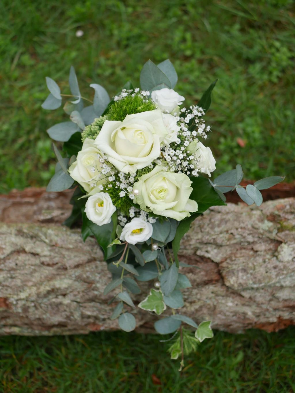 Ein Brautstrauß aus weißen Rosen, Schleierkraut und Grün, der auf einem Baumstamm liegt und von grünem Gras umgeben ist.