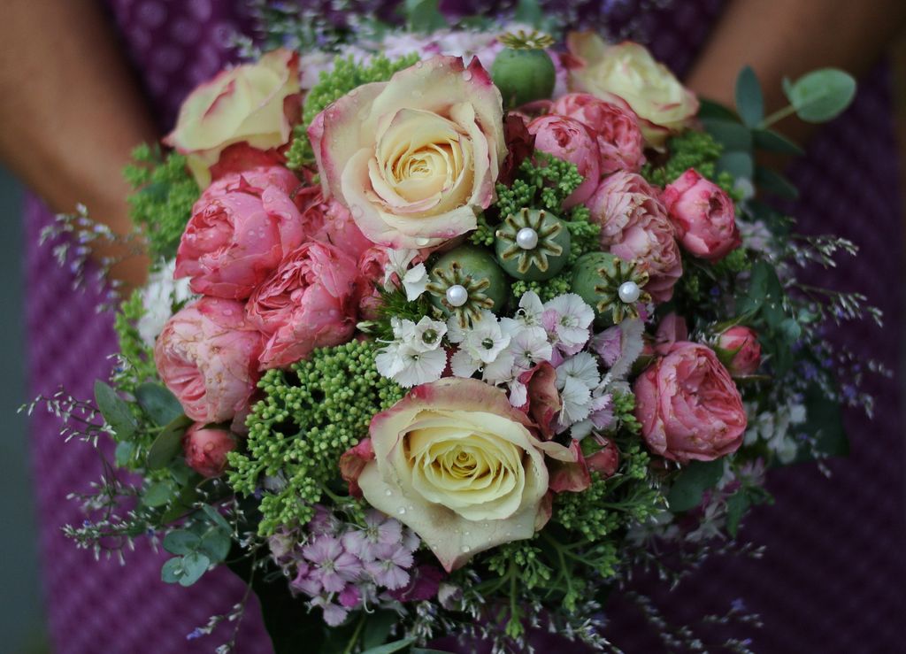 Nahaufnahme eines Brautstraußes mit rosa und weißen Rosen, grünen Samenkapseln, kleinen weißen und rosa Blüten, gehalten vor einem violetten Kleid.