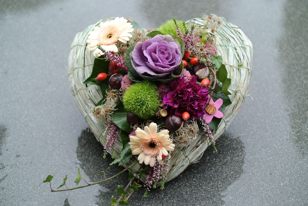 Herzförmiges Gesteck aus verschiedenen Blumen wie lila Zierkohl, weißen Gerbera, pinkfarbener Nelke und grünen Moosballen auf einer grau glänzenden Oberfläche.