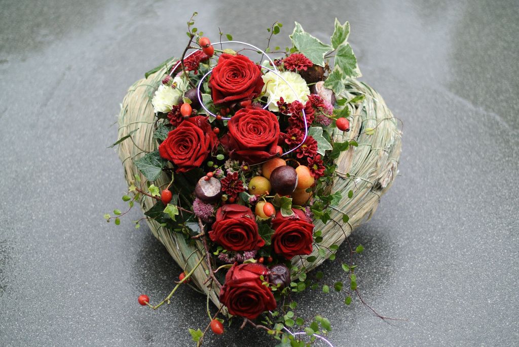 Ein herzförmiges Gesteck aus getrocknetem Gras, dekoriert mit roten Rosen, kleinen roten Beeren, grünen Blättern und weiteren Blumen, auf einem grauen Untergrund.