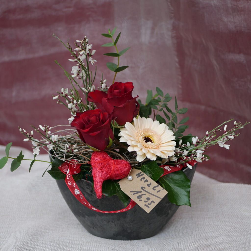 Ein Blumenarrangement in einer schwarzen Vase mit roten Rosen, einer weißen Gerbera, grünen Blättern, kleinen weißen Blüten und einem roten Herzanhänger mit einer Preiskarte.