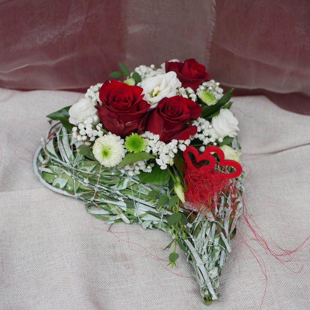 Herzförmiges Blumenarrangement mit roten Rosen, weißen Blumen und grünen Blättern auf einem hellen Stoffuntergrund.