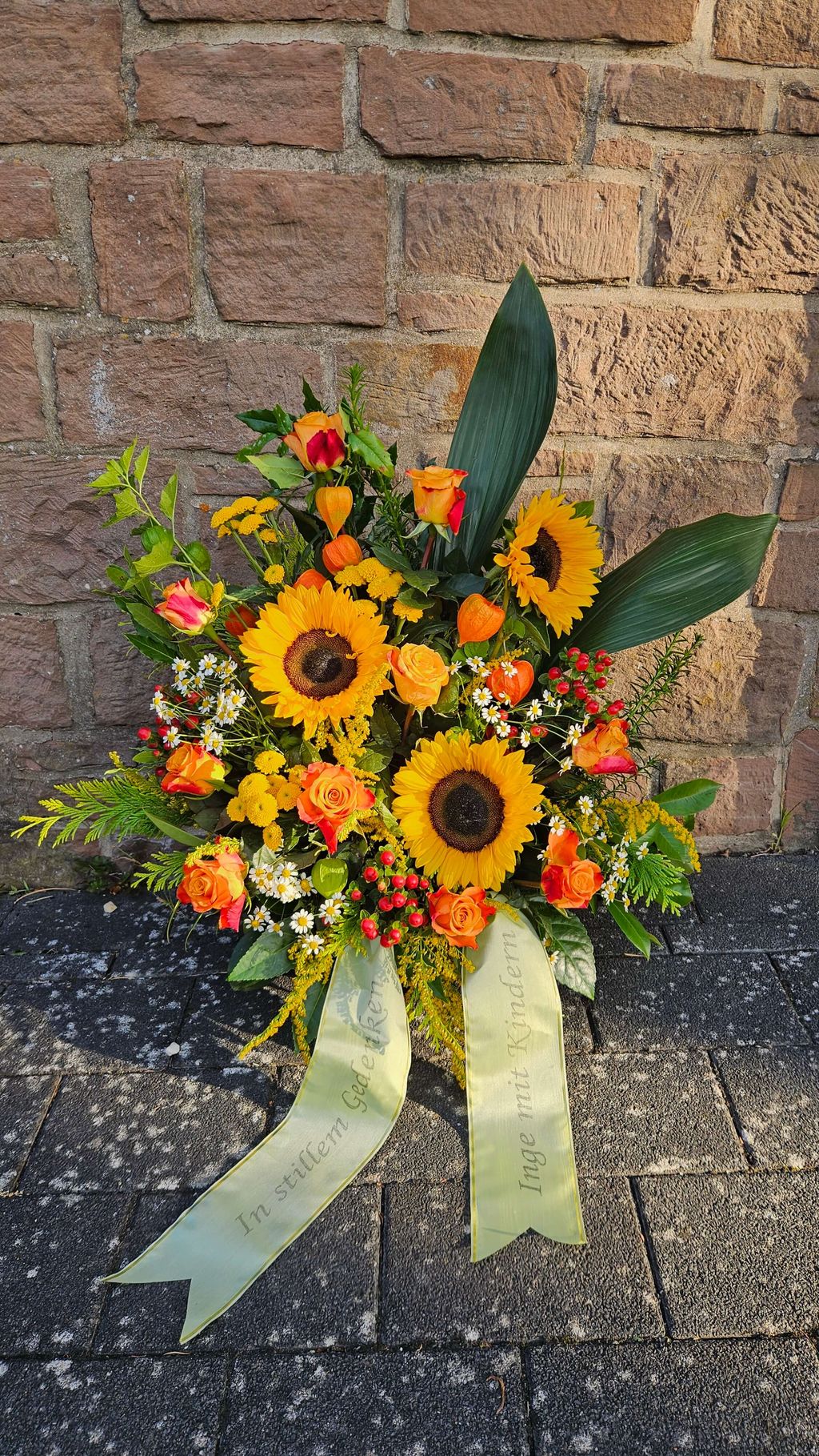 Bunte Trauerblumenarrangement mit Sonnenblumen, Rosen und anderen Blumen vor einer Steinmauer auf einem gepflasterten Boden mit zwei Schleifenbandern.