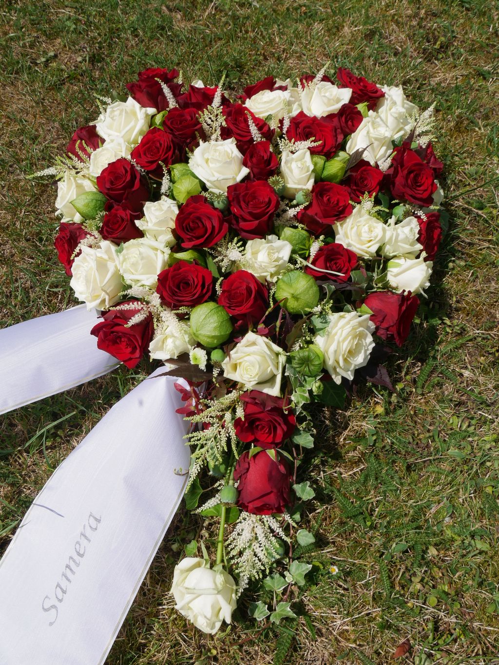 Herzförmiges Blumenarrangement aus roten und weißen Rosen mit weißen Schleifen auf Gras, kennzeichnend für Trauerfloristik.