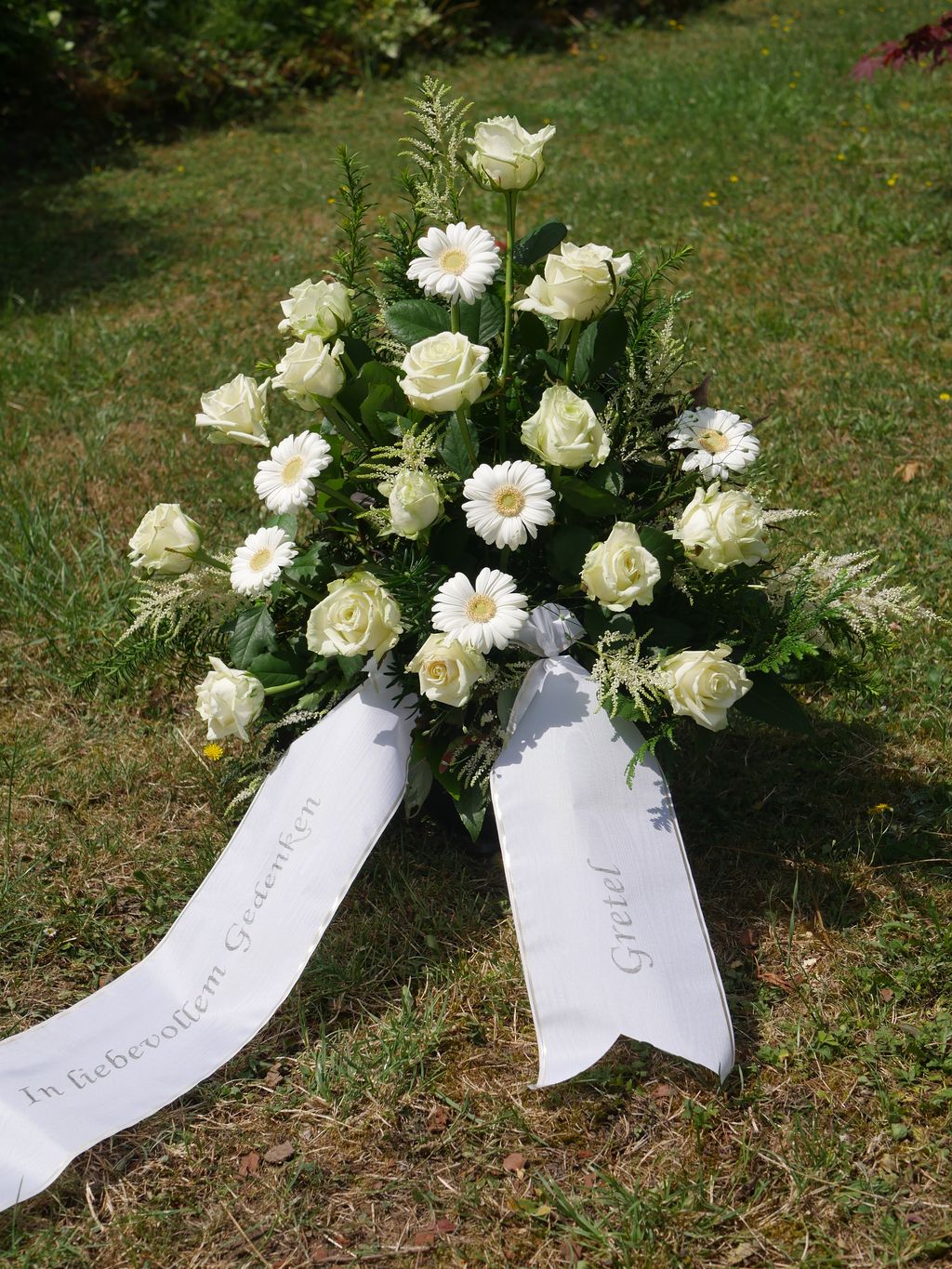Strauß aus weißen Rosen und Gerbera mit zwei weißen Schleifen auf einer Rasenfläche, eine Schleife trägt die Aufschrift "In liebevollem Gedenken", die andere den Namen "Gretel".