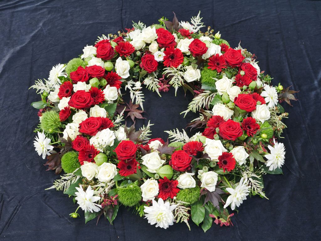 Trauerkranz aus roten Rosen, weißen Rosen, roten Gerberas, grünen Chrysanthemen und weiteren weißen Blumen auf schwarzem Hintergrund.