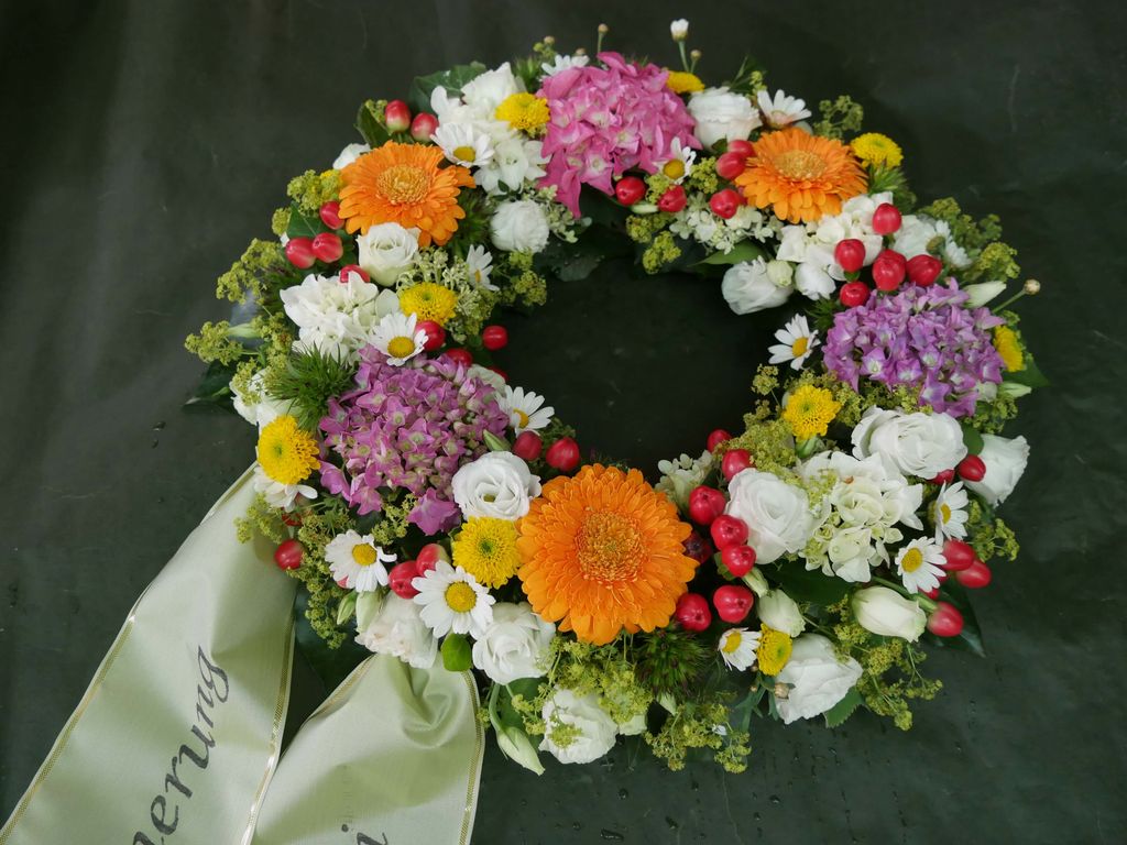 Bunter Trauerkranz aus verschiedenen Blumen wie weißen Rosen, orangefarbenen Gerbera, lila Hortensien, roten Beeren und weißen Gänseblümchen, mit einem hellgrünen Schleifenband.