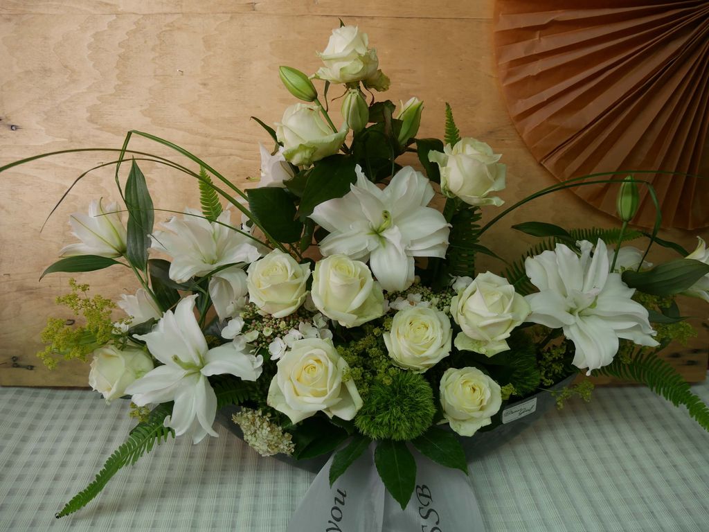 Strauß aus weißen Rosen, Lilien und grünen Blättern in einer Trauergesteck-Anordnung auf kariertem Tuch vor Holzwand und braunem Papierfächer.
