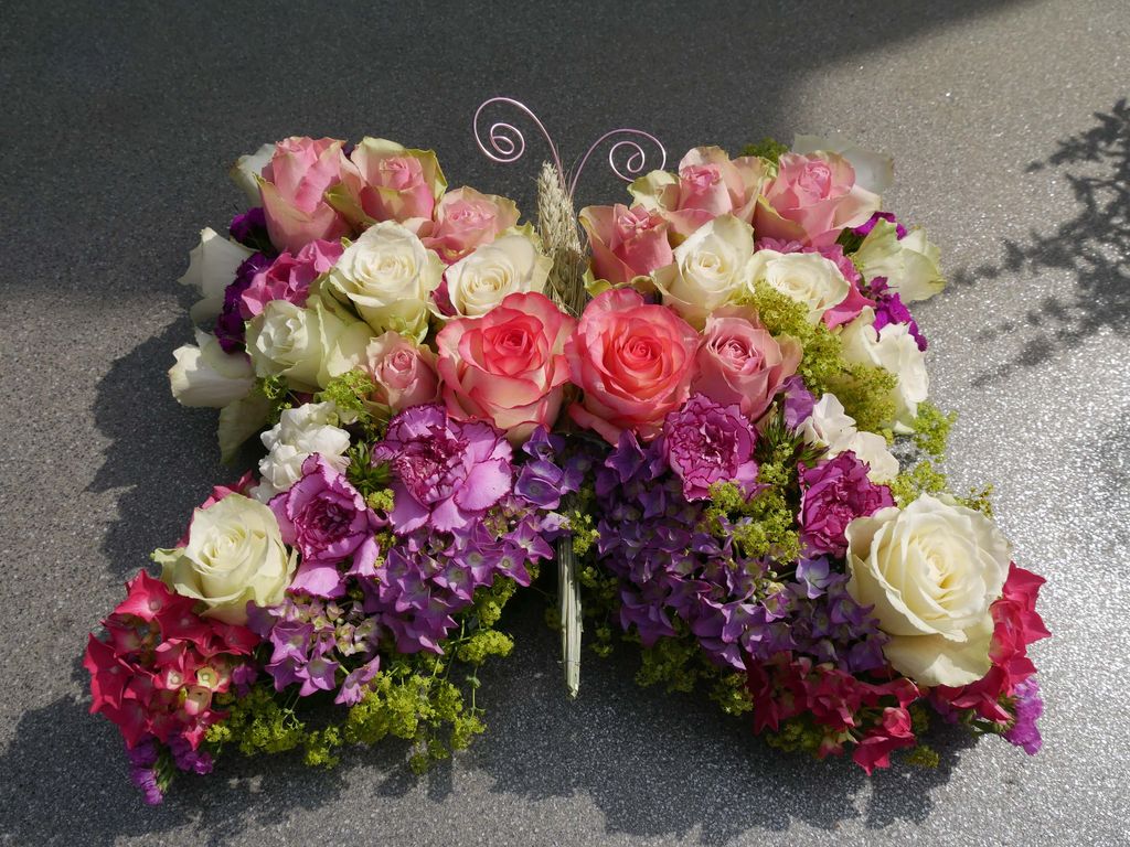 Schmetterlingsförmiges Blumenarrangement mit Rosen in Weiß, Rosa und Rot sowie lila und grünen Akzenten auf einem grauen Untergrund.