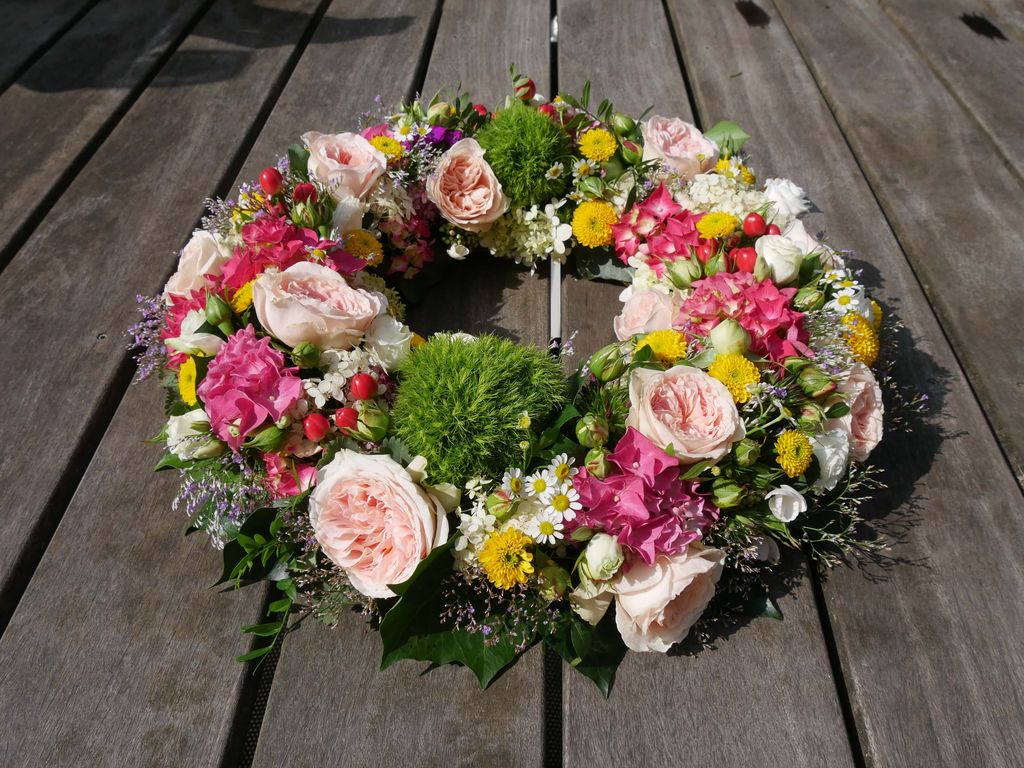 Bunter Blumenschmuck in Form eines Kranzes mit rosa Rosen, gelben und weißen Blüten sowie grünen Blättern auf einer Holzunterlage.