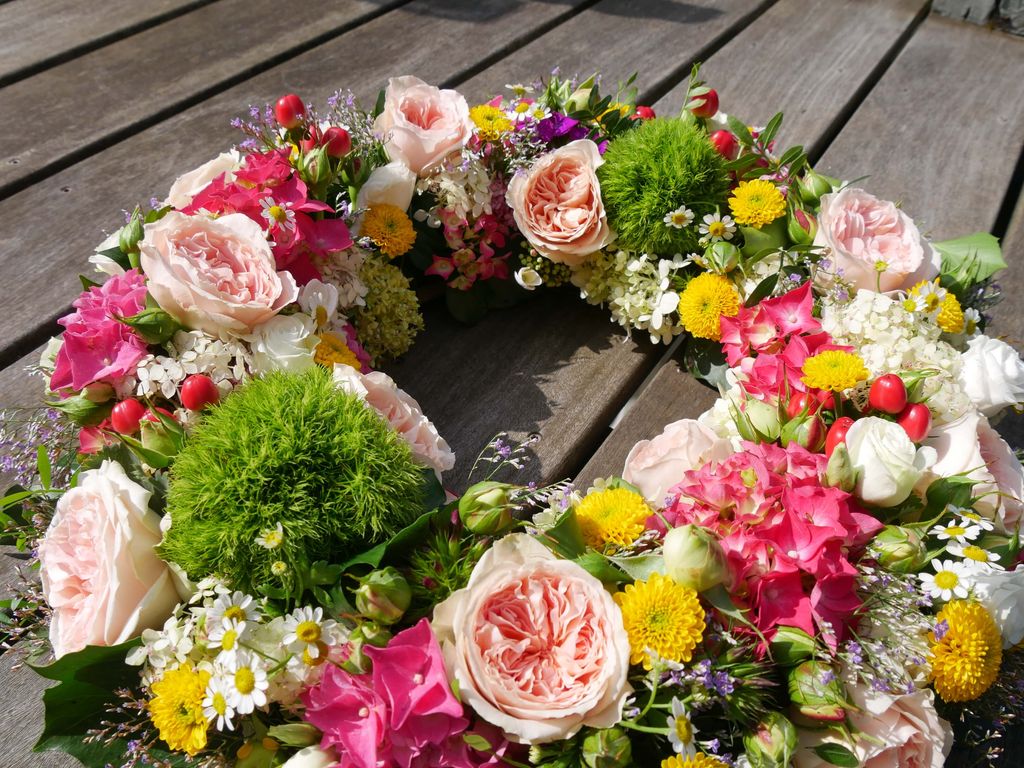 Bunter Trauerkranz aus verschiedenen Blumen wie Rosen, Gänseblümchen, gelben Blüten und grünen Moosen auf Holzdielen.