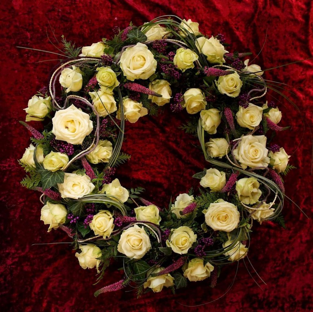 Trauerkranz aus cremefarbenen Rosen und grünen Blättern auf rotem Samthintergrund.