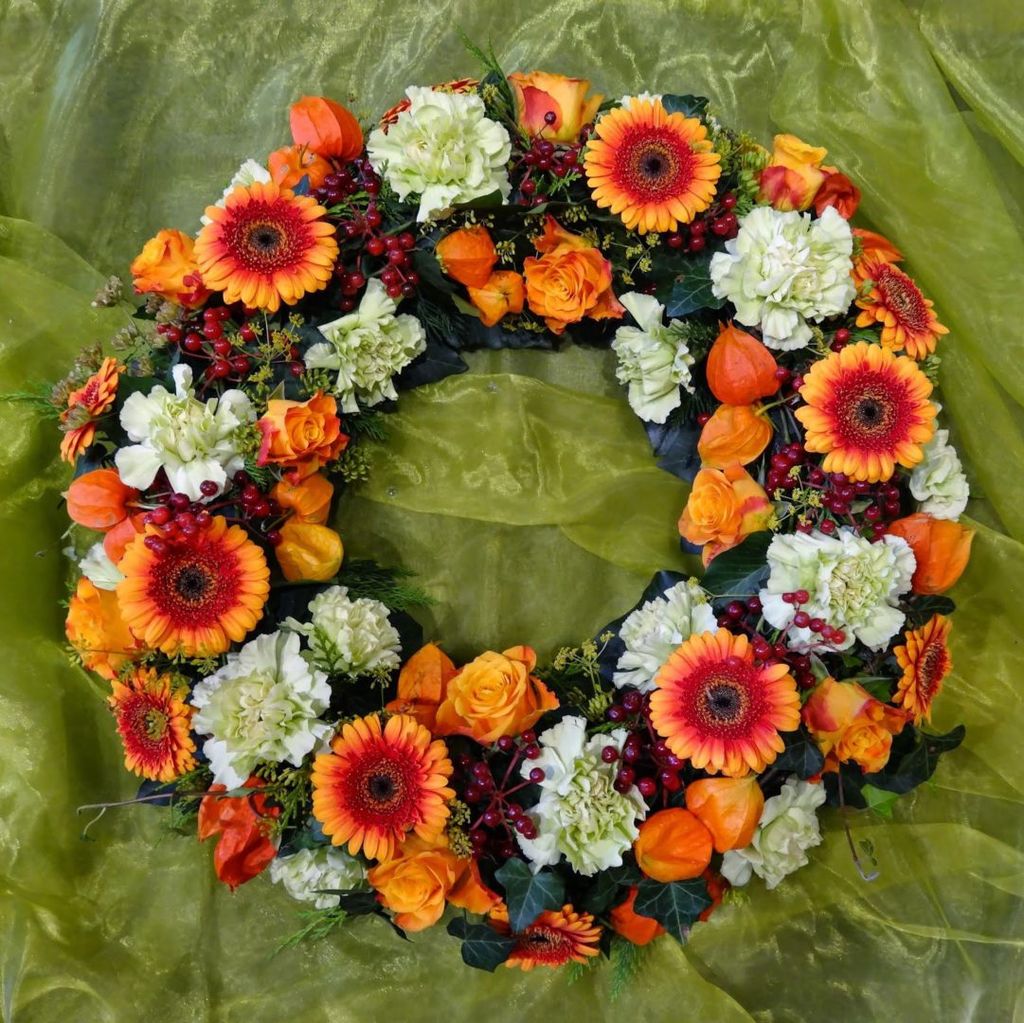 Bunter Trauerkranz aus orangen Gerbera, Rosen, weißen Nelken und roten Beeren auf grünem Stoffliegefläche.