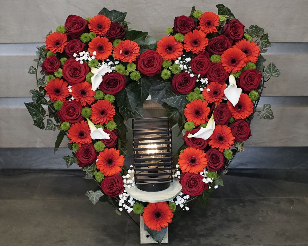 Herzförmiges Blumenarrangement aus roten Rosen, orangefarbenen Gerbera, weißen Lilien und grünen Blättern mit einer beleuchteten Lampe in der Mitte, auf einem dunklen Boden vor einer grauen Wand.