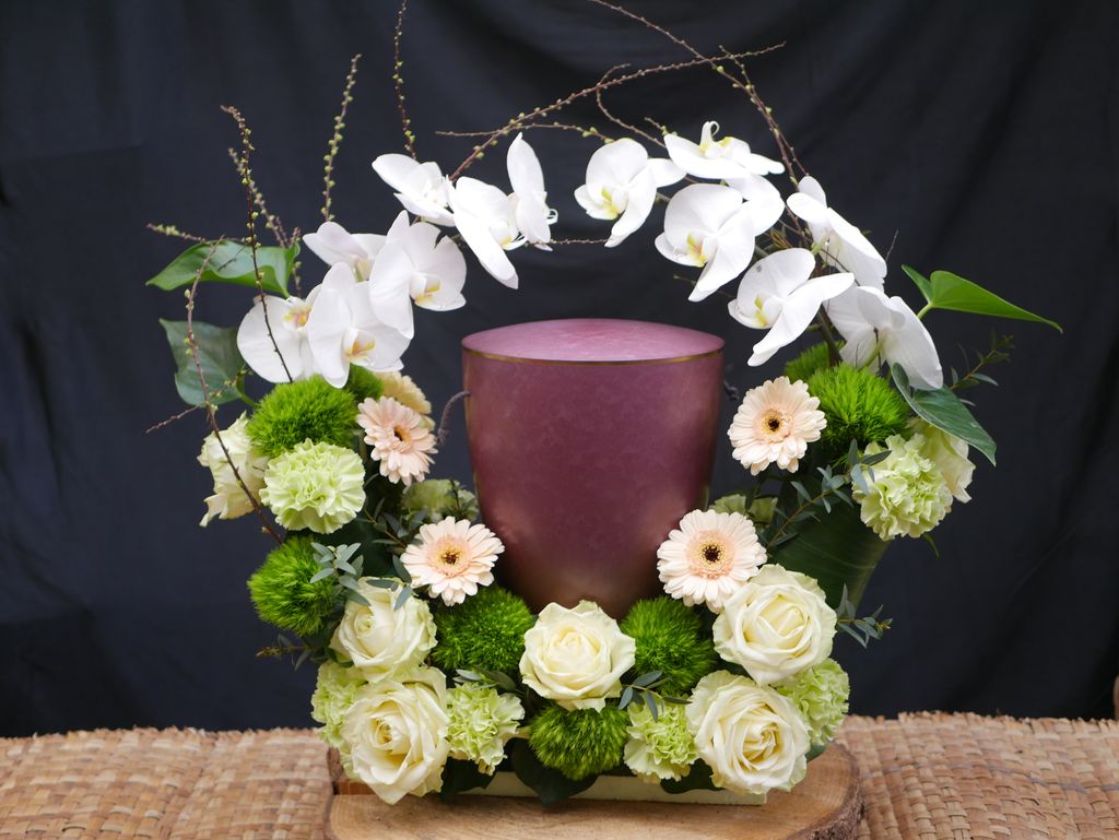 Ein Blumenarrangement mit weißen Orchideen, weißen Rosen und grünen Akzenten um eine violette Urne vor einem schwarzen Hintergrund.