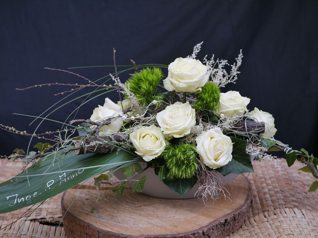 Ein Blumenarrangement mit weißen Rosen, grünen Moosbällen und verschiedenen Zweigen in einer weißen Schale auf einer Holzscheibe vor einem dunklen Hintergrund.
