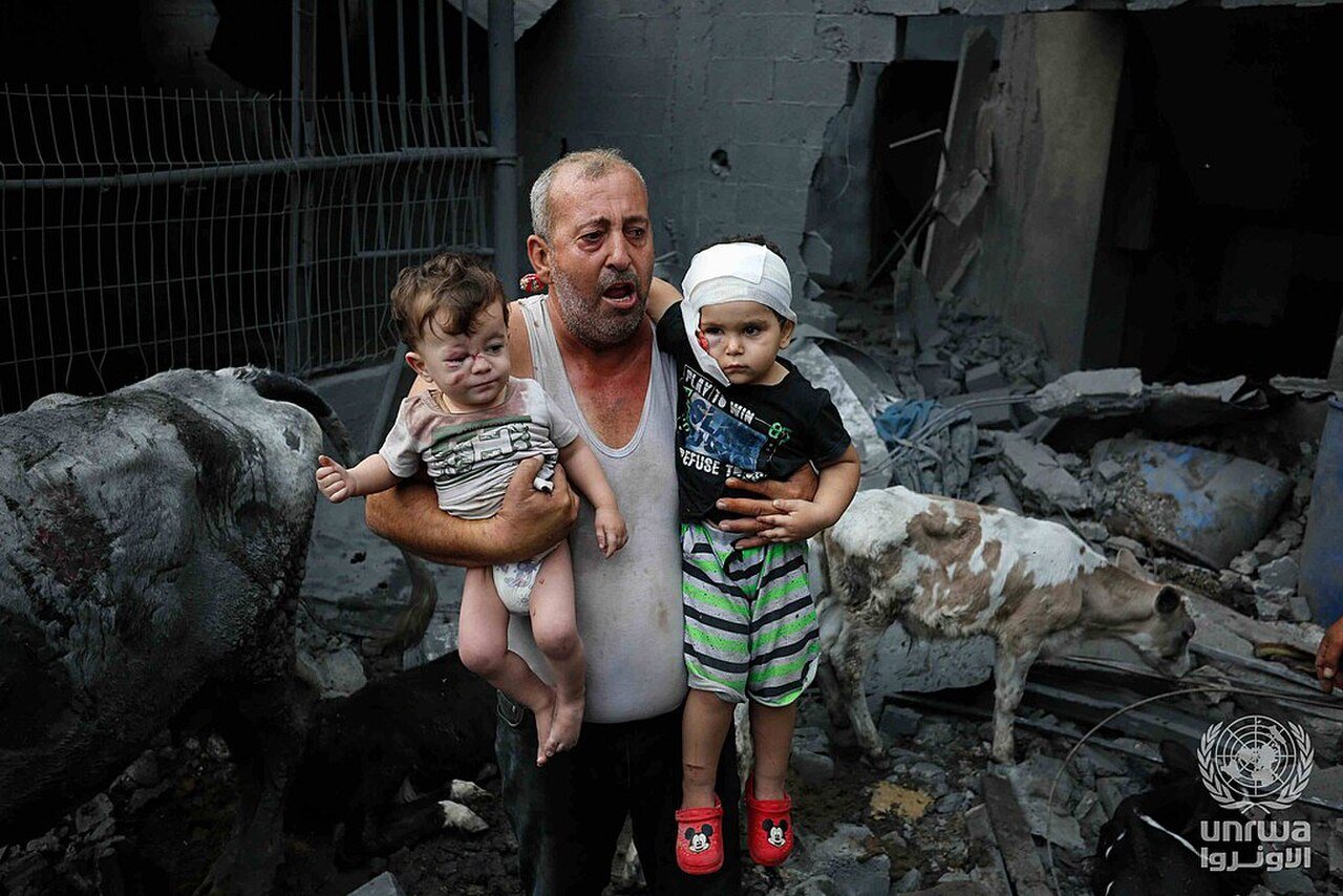 A Palestinian refugee carries his injured grandchildren from the Israeli bombing of Nuseirat Camp, Gaza Strip