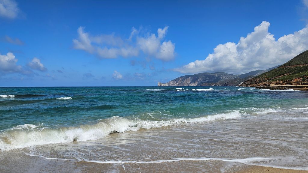Waves rolling onto a sandy beach with a partly cloudy blue sky and rocky coastline in the background.