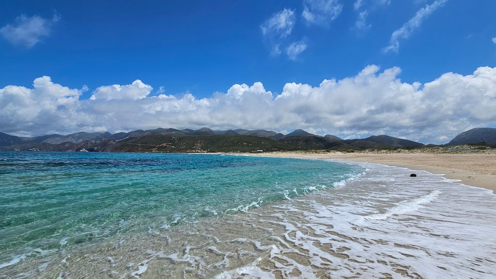 Clear ocean with turquoise water meeting a wide sandy beach under a blue sky with white clouds, with hills and mountains in the background.