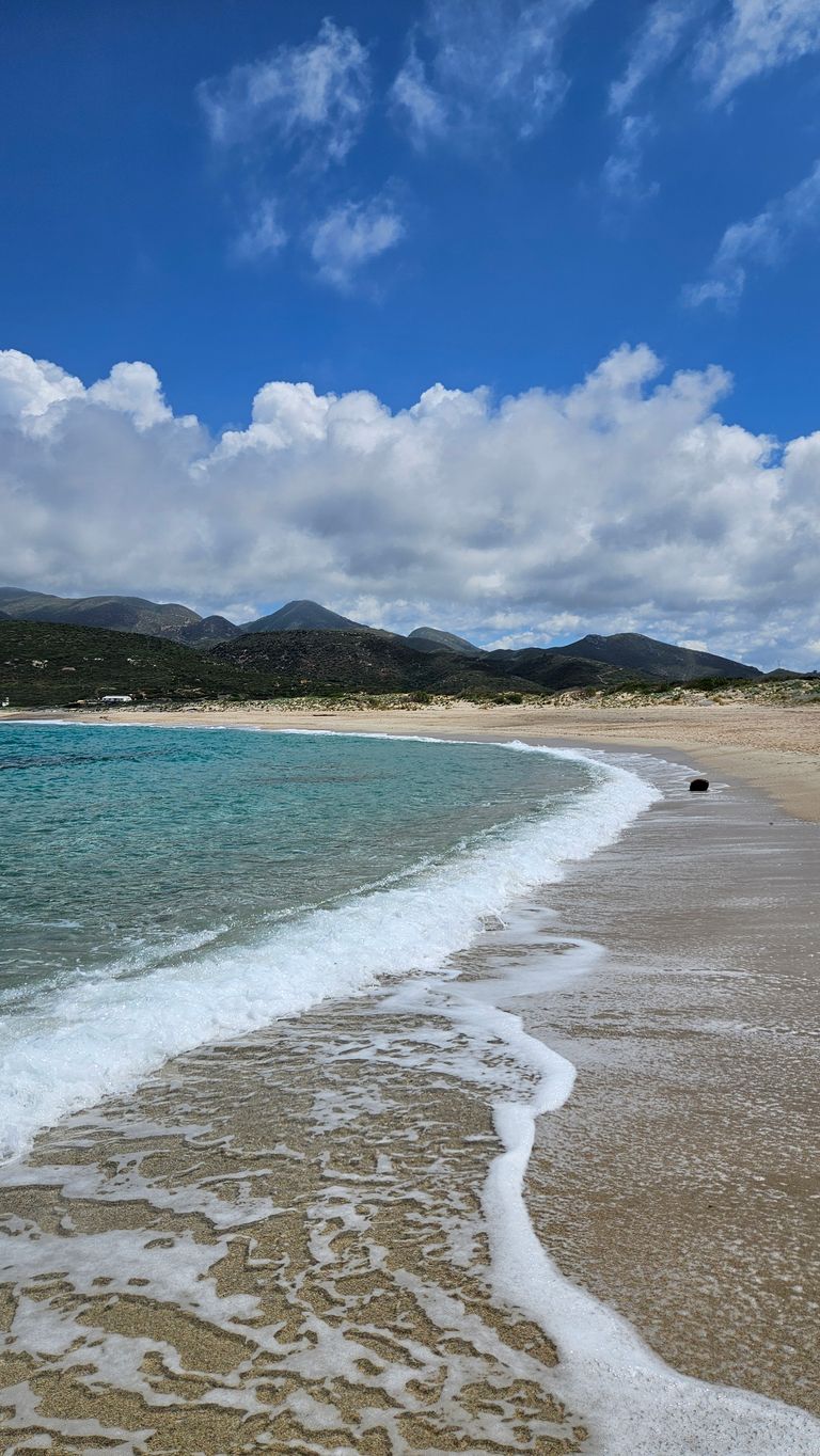Gentle waves hitting the sandy beach with turquoise water and a mountain landscape in the background under a partly cloudy blue sky.