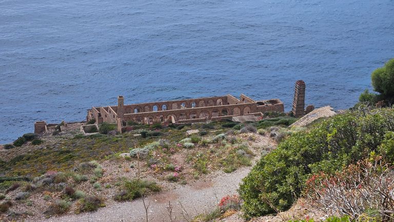 Ruins of an old rectangular stone structure with arched windows by the coastline overlooking the sea, with grassy and shrub-covered hills in the foreground.