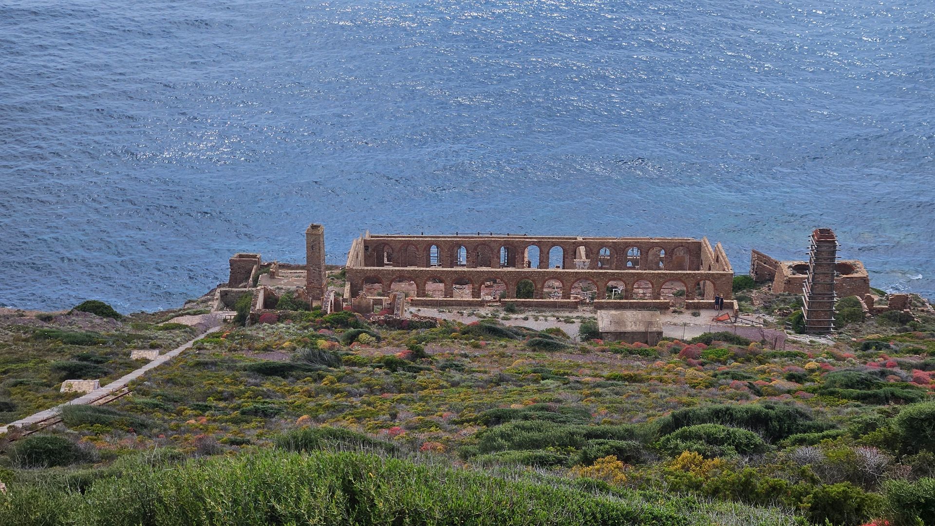 Ruins of a stone wall with arches located on the shore of a large body of water, surrounded by green and flowering shrubs on a hill.