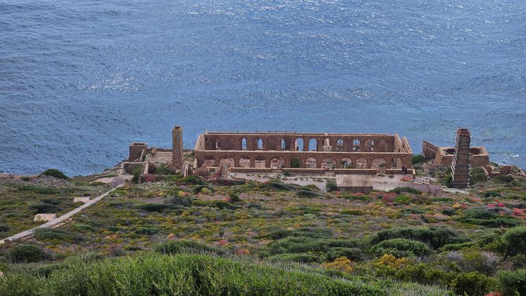 Ruins of a stone wall with arches located on the shore of a large body of water, surrounded by green and flowering shrubs on a hill.