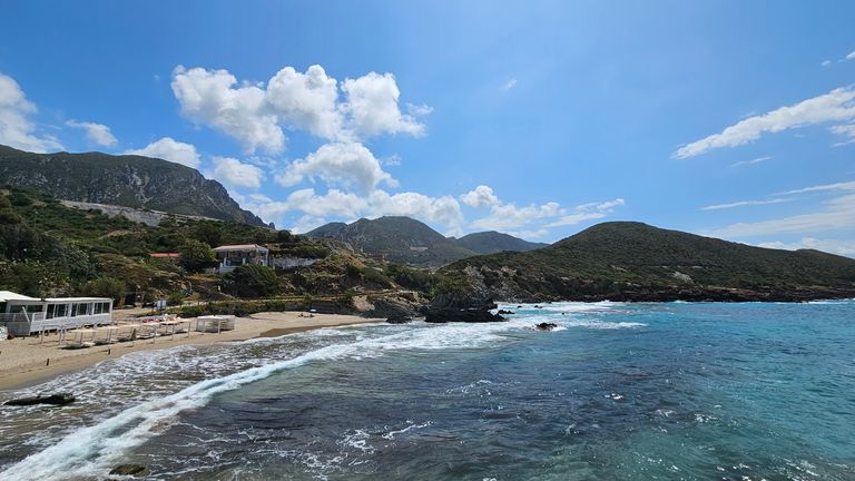 Coastal landscape with a rocky beach, blue sea, and wooded hills under a bright blue sky with some clouds.