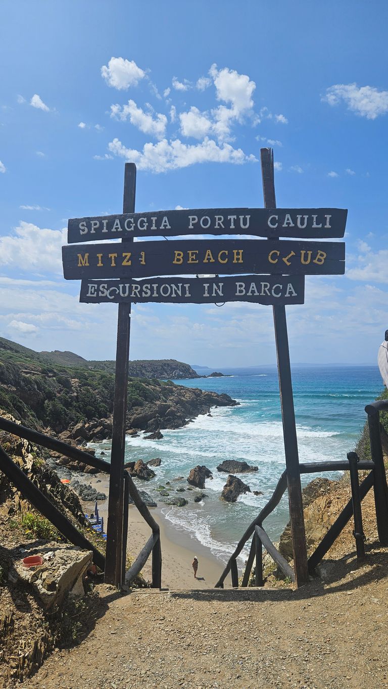 Entrance to Spiaggia Portu Cauli beach with a wooden sign and stairs leading down to a rocky beach with blue sea and partly cloudy sky.