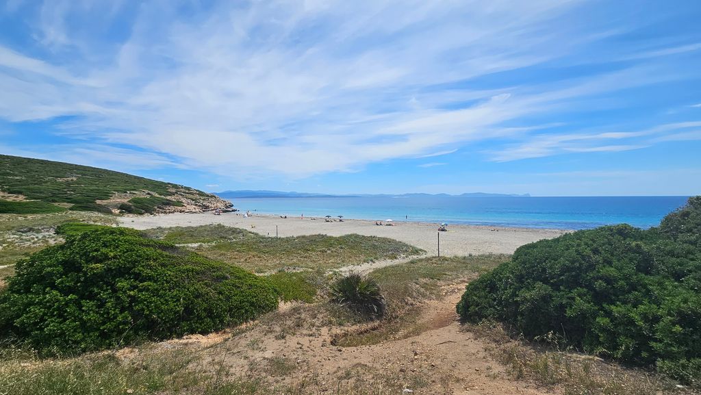 A wide sandy beach with a few umbrellas and people, surrounded by green hills under a bright blue sky with scattered clouds.