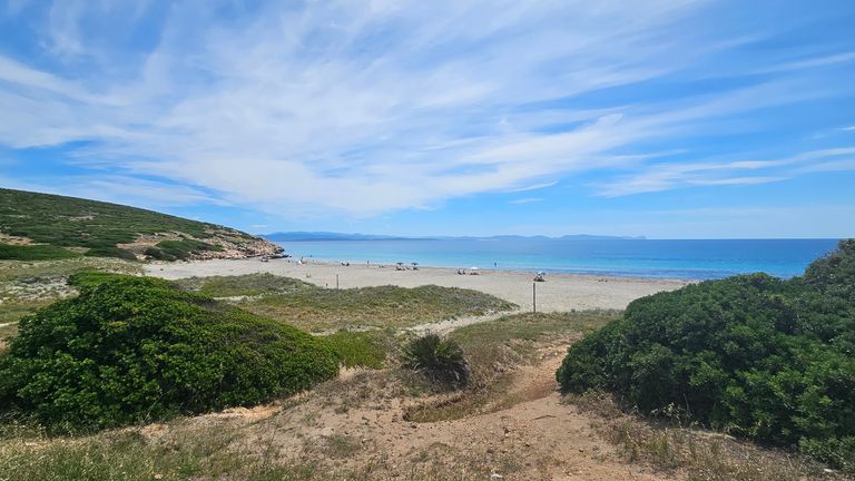 A wide sandy beach with a few umbrellas and people, surrounded by green hills under a bright blue sky with scattered clouds.
