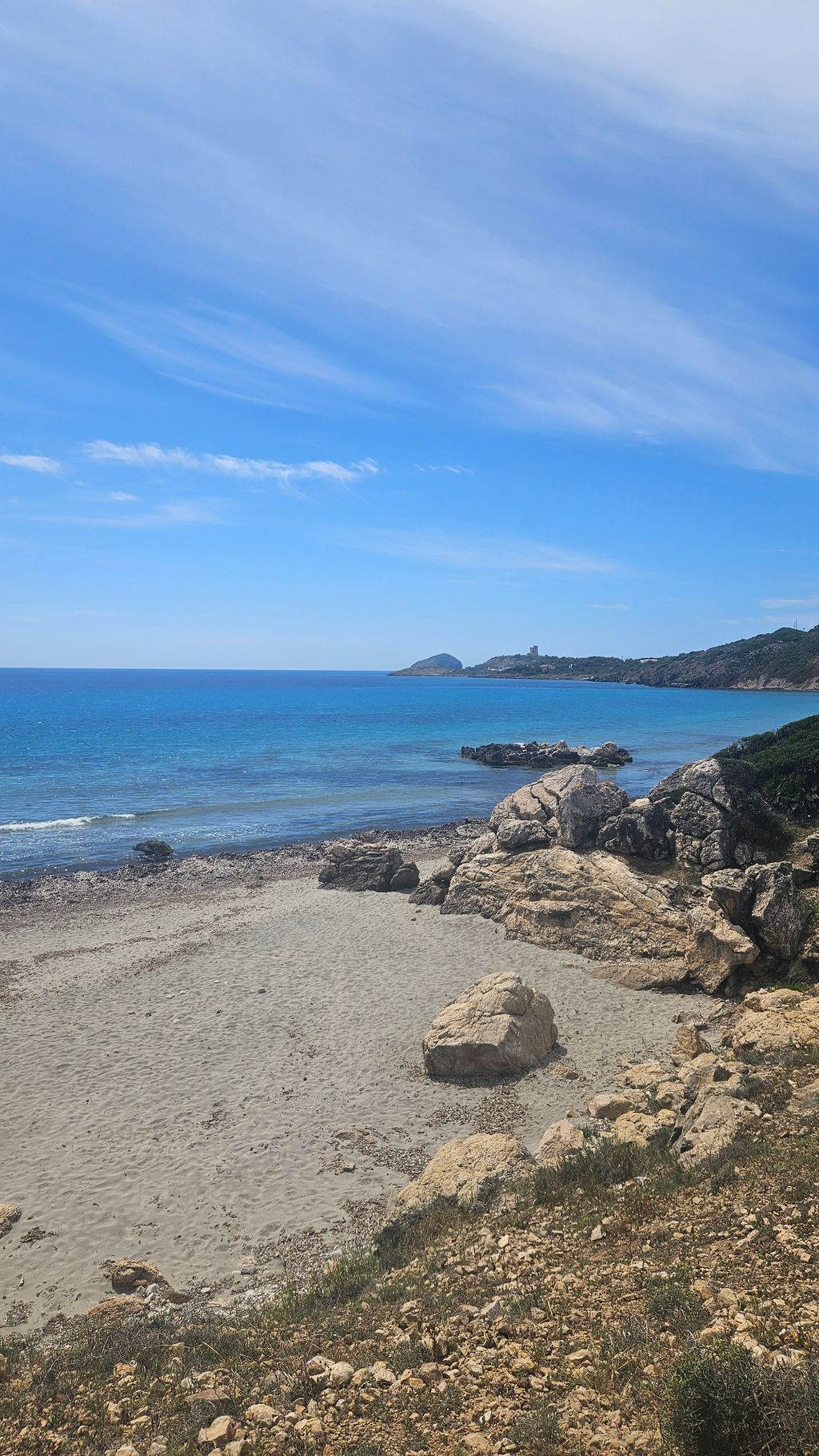 Pebble and sandy beach with rocks along the coastline and view of the blue sea under a partly cloudy sky.