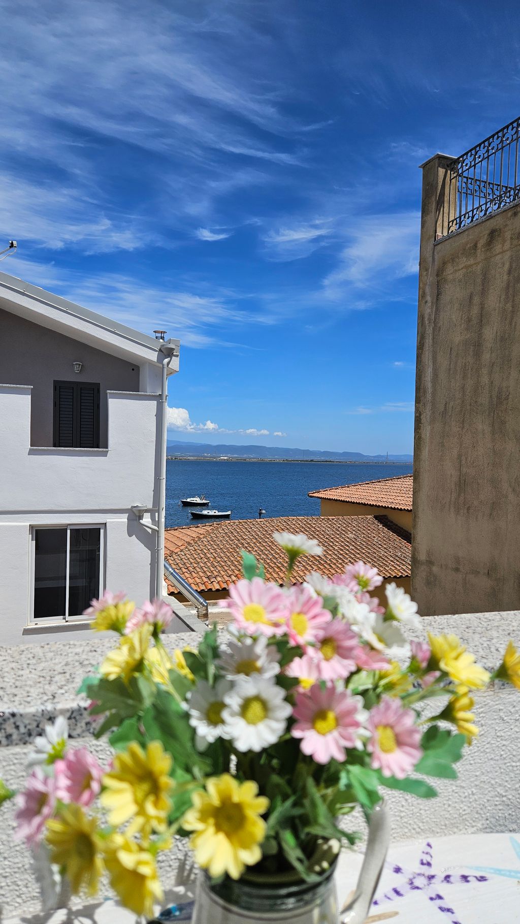 View of the sea with a boat, visible buildings with red tile roofs and a white house, with a blurred bouquet of yellow, white, and pink flowers in the foreground.