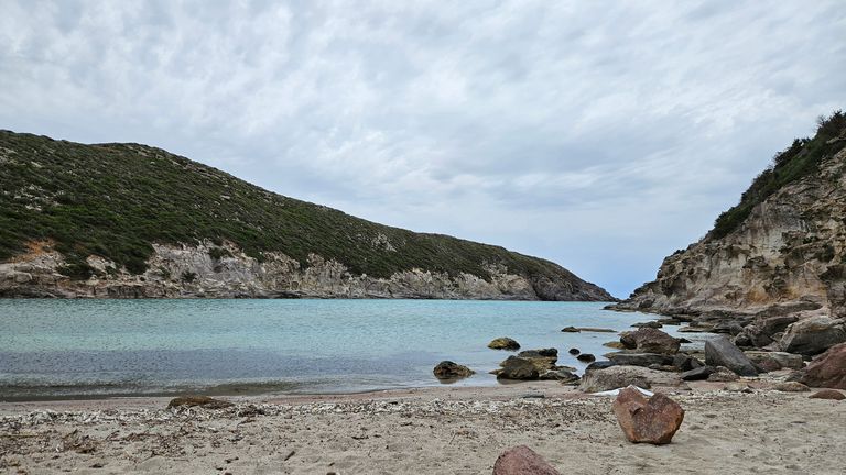 Beach with sand and rocks in the foreground, calm turquoise sea surrounded by green-covered cliffs under a cloudy sky.