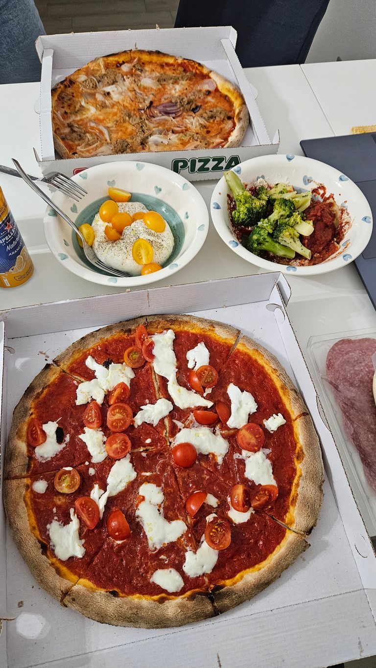 Two pizzas in boxes on a white table, accompanied by two bowls with sides: one with broccoli and tomato sauce, the other with burrata and yellow cherry tomatoes. Also visible are a can of drink and a package of sausage.