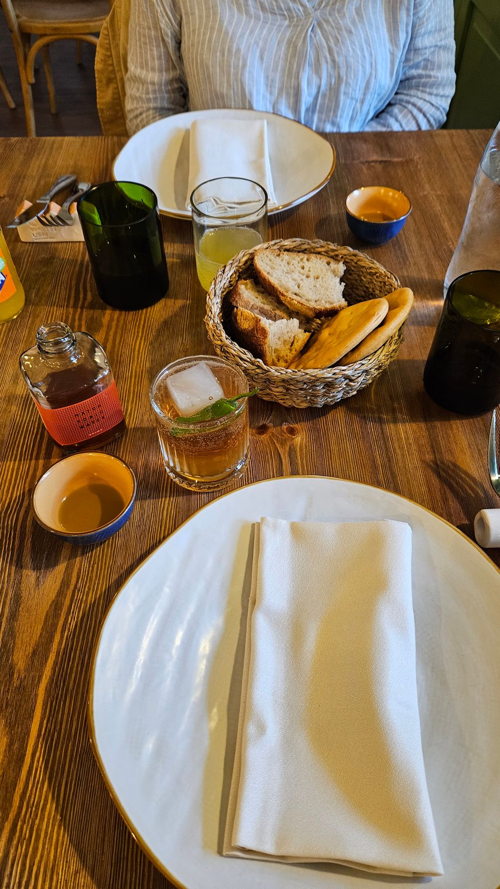View of a set wooden table in a restaurant with two white plates, folded napkins, a bread basket containing two types of bread, two glasses with drinks, a small bottle and small bowls.