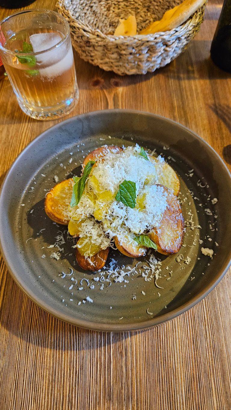Fried potato slices on a gray plate, garnished with grated cheese and fresh mint leaves, placed on a wooden table. In the background, a glass of drink with ice and mint and a woven basket with bread.