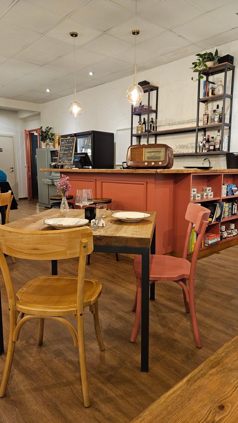 A cozy restaurant or café with a wooden table, two chairs, set plates, and a vase with flowers. In the background, there is a red bar counter with an old radio and shelves holding bottles and glasses.