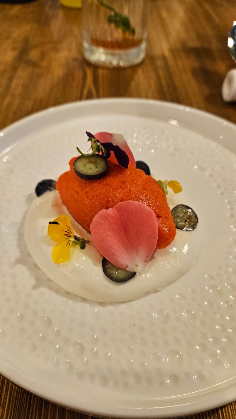 An elegant dish featuring an orange mousse on a white sauce, garnished with small blueberry slices and edible flowers, served on a white plate with a dotted texture.