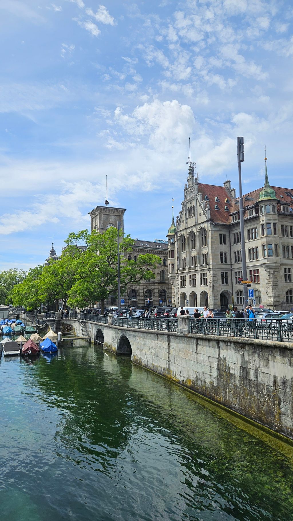 Riverside street with historic buildings, moored boats, and people along the railing under a partly cloudy sky.